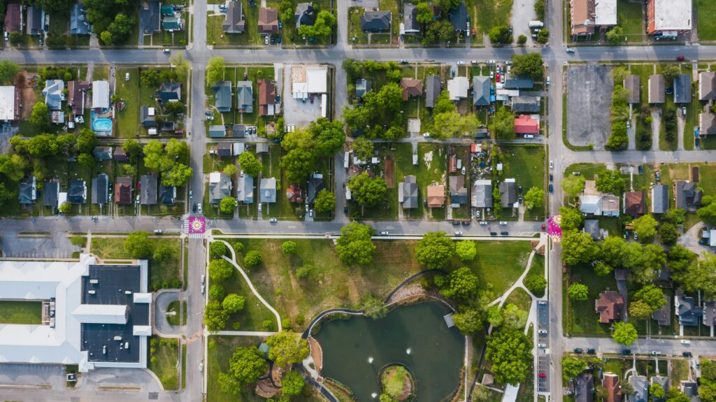 High-angle shot of a suburban neighborhood with streets, houses, and a park with a pond, showcasing vibrant greenery.